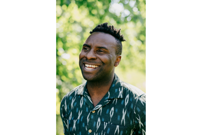 Jimi Famurewa standing outdoors, smiling confidently, dressed casually, with a background of greenery.